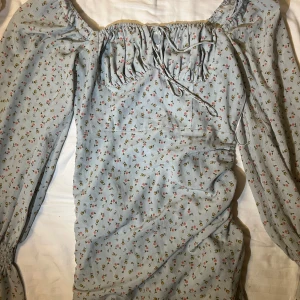 Blommig klänning från ADOORE - Charmig ljusblå klänning från ADOORE med ett sött blommönster i rosa och grönt. Klänningen har långa ärmar och en smickrande offshoulder-design med knytning framtill. Perfekt för en romantisk look! Går att justera längden på genom dragsko.