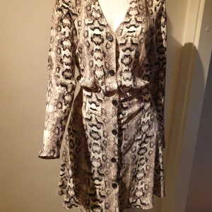 Ormmönstrad klänning med knappar - Snygg klänning med ormmönster och knappar framtill. Den har en v-ringad hals och långa ärmar. Perfekt för en trendig look.