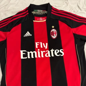 AC Milan fotbollströja från Adidas - Snygg AC Milan tröja i rött och svart med klassiska ränder. Adidas-logga och Fly Emirates tryck på framsidan. Perfekt för fans av laget! Då de är en äldre tröja blir den lite dyrare men priset går att diskutera!