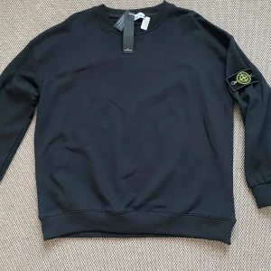 Svart sweatshirt från Stone Island - Säljer en stilren svart sweatshirt från Stone Island med deras ikoniska logga på ärmen. Tröjan har en klassisk rund hals och långa ärmar. Perfekt för en avslappnad och trendig look. Har inte använd den, fick den i en present på mitt födelsedag. Säljer för att den har för liten 