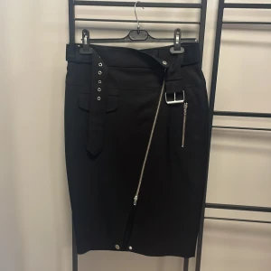 Svart kjol från Jean Paul Gaultier - Snygg svart kjol från Jean Paul Gaultier med coola detaljer som dragkedja och bälte. Kjolen har en stilren design med en modern twist och passar perfekt för den som vill sticka ut. Storlek EUR 40.