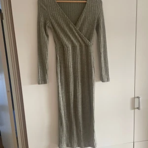 Grå ribbad omlottklänning - Snygg grå/grön långklänning med ribbat mönster och omlottdesign. Klänningen har långa ärmar och en v-ringad halslinje, perfekt för en stilren look. Den är både bekväm och trendig, idealisk för kyligare dagar.