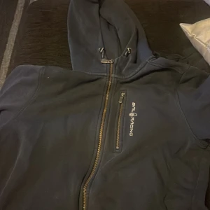 Mörkblå hoodie från Sail Racing - Säljer en mörkblå hoodie från Sail Racing med dragkedja och huva. Den har en broderad logga på bröstet och är tillverkad i ett mjukt material som känns skönt mot huden. Perfekt för kyliga dagar.