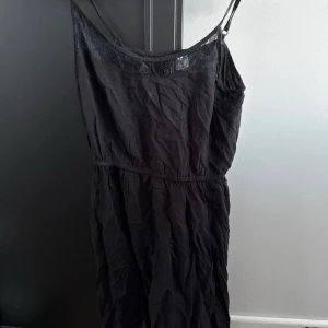 Svart klänning med spetsdetaljer - Snygg svart klänning med smala axelband och spetsdetaljer vid halsringningen. Perfekt för en stilren look. Klänningen har en enkel och elegant design.