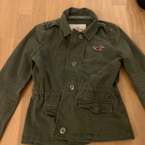Grön jacka från Hollister - Säljer en grön jacka från Hollister med knappar och en broderad logga på bröstet. Jackan har en justerbar midja med knytband och långa ärmar. Super fin!!
