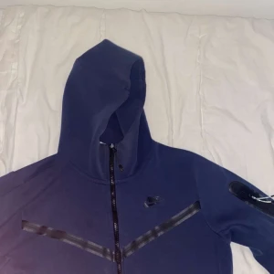 Mörkblå hoodie från Nike - Säljer en mörkblå hoodie från Nike med dragkedja och huva. Jackan har en stilren design med diskreta detaljer och en ficka på ärmen. Perfekt för en sportig look.