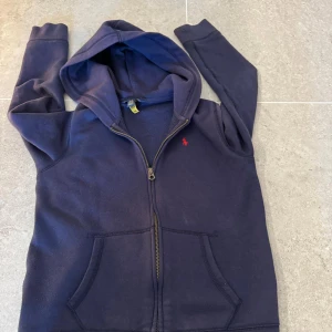 Blå zip hoodie från Ralph Lauren - Säljer en snygg blå hoodie från Ralph Lauren med dragkedja och broderad röd logga på bröstet. Tröjan har en mysig huva och praktiska fickor framtill.  Tveka inte på att fråga! 