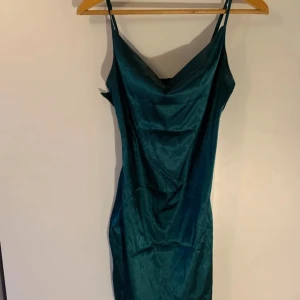 Mörkgrön sidenklänning - Elegant mörkgrön klänning i siden med smala axelband och en smickrande passform. Perfekt för en stilfull kväll med sin glänsande yta och enkla design. Klänningen har en vacker drapering framtill som ger en extra touch av elegans.