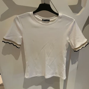 Zara tshirt med spets - Snygg vit topp från Zara med korta ärmar och dekorativa spetsdetaljer i svart och guld vid ärmsluten. Helt oanvänd och är slutsåld på hemsidan❣️