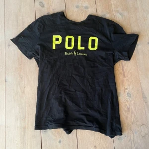 Svart t-shirt från Ralph Lauren - Säljer en svart t-shirt från Ralph Lauren med stor gul text 'POLO' på framsidan. T-shirten har korta ärmar och är perfekt för en avslappnad stil.