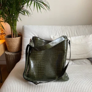 Grön axelväska i krokodilmönster - Snygg grön axelväska med krokodilmönster. Väskan har en justerbar axelrem och ett rymligt huvudfack med dragkedja. Perfekt för att bära med sig alla nödvändigheter med stil.