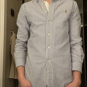 Randig skjorta från Ralph Lauren - Säljer en stilren ljusblå och vit randig skjorta från Ralph Lauren. Skjortan har en klassisk krage och knappar framtill. Den är broderad med den ikoniska loggan på bröstet och är tillverkad i 100% bomull för en bekväm passform. Ny pris 1500, priset kan diskuteras och om ni har frågor är det bara att höra av er! 