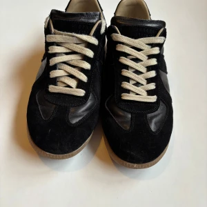 Svarta Margiela sneakers - Snygga svarta sneakers från Maison Margiela med vita snören och en klassisk design. Skorna har en kombination av skinn och mocka, vilket ger en stilren look. Perfekta för en avslappnad stil. Inga defekter. Använd lite. Storlek 42 men passar 42/5-43