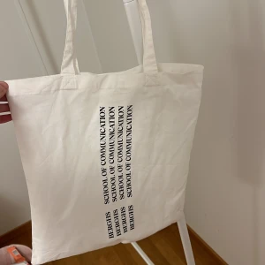 Vit tygväska med text - Säljer en vit tygväska med texten 'BERGHS SCHOOL OF COMMUNICATION' tryckt i svart. Perfekt för att bära böcker eller andra småsaker. Väskan har två långa handtag för enkel bärning över axeln.