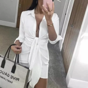 Zara S/XS Vit klänning med knytning - Snygg vit klänning med knytning framtill och långa ärmar. Klänningen har en djup v-ringning och är i en kort modell. Går att knyta med rosett eller bara löst. Fint skick! Strl S men liten i strl. 