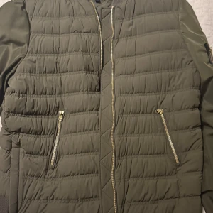 Olivgrön quiltad jacka - Säljer en olivgrön quiltad jacka med dragkedja framtill och på fickorna. Jackan har en stilren design med långärmade ärmar och en praktisk ficka på ärmen. Perfekt för vår och höstdagar. Skicket är 10/10