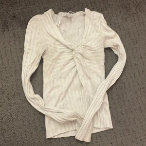 Vit ribbad topp med knyt - Säljer en stilren vit ribbad topp med knyt framtill. Toppen har långa ärmar och en v-ringad design som ger en elegant look. Perfekt för att matcha med både jeans och kjol. Hör av dig vid intresse!