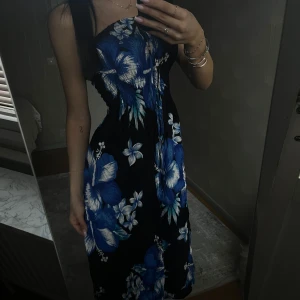 Blommig axelbandslös klänning - Snygg axelbandslös klänning med ett tropiskt blommönster i blått och vitt på en svart bakgrund. Klänningen har en slits men det ser ändå bra ut och som ger en extra touch av stil! Perfekt för sommarens varma dagar!