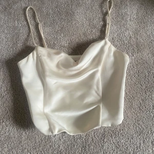 Vit satinlinne från Zara - Elegant vit satinlinne med tunna axelband och draperad halsringning. Perfekt för en stilren look. Superfin korsett liknande linne från Zara i satin. Säljer då den är för liten men älskar toppens passform. Skriv för fler bilder och frågor!💕