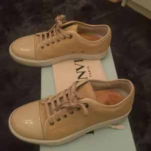 Beige sneakers från Lanvin - Snygga beige sneakers från Lanvin med snörning och en stilren design. Skorna har en blank yta på tån och en vit sula som ger en modern touch. Perfekta för en avslappnad men elegant stil. Det är helt nya. Jag har sula i så den tar jag bort om nån vill köpa. Allt ingår box osv. Perfekt inför sommaren. Storlek 39 men passar 40