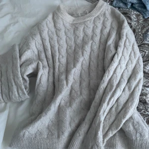 Flätad tjocktröja - Mysig beige/vit stickad tröja med kabelstickat mönster. Perfekt för kyliga dagar med sin varma och bekväma design. Tröjan har långa ärmar och ribbade kanter vid hals, ärmslut och nederkant. 