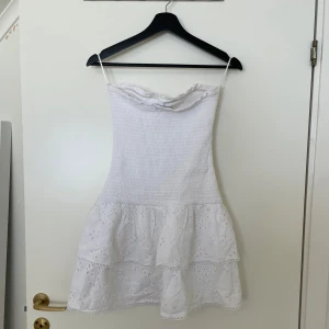 Vit sommarklänning med spetsdetaljer - Charmig vit klänning, inga axelband och vacker spetsdetalj på kjolen. Klänningen har en smockad överdel som ger en bekväm passform. Oanvänd.