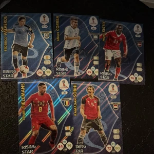 Fotbollskort från FIFA World Cup 2018 - Samling av fem fotbollskort från Panini, FIFA World Cup 2018. Kort med spelare som Federico Valverde, Ángel Correa, Pione Sisto, Youri Tielemans och Ramadan Sobhi. Varje kort har spelarinformation och 'Rising Star'-märkning. Perfekt för samlare av fotbollsmemorabilia.
