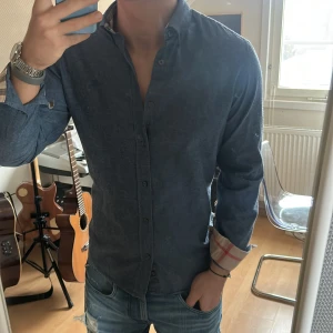 Burberry skjorta - Snygg blå skjorta från Burberry Brit med klassiskt rutigt mönster på insidan av ärmsluten. Skjortan har långa ärmar och knappar framtill. Perfekt för en stilren look.