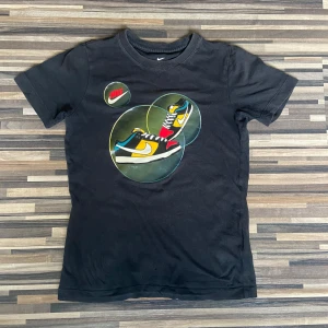 Svart t-shirt från Nike med grafiskt tryck - Snygg svart t-shirt från Nike med ett coolt grafiskt tryck på framsidan. Perfekt för en sportig look. Tillverkad i mjukt material för extra komfort.