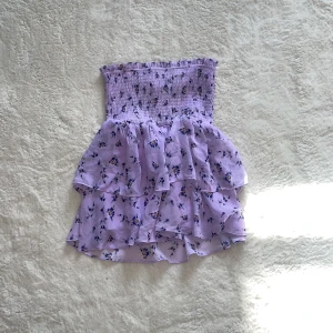 Lila blommig tubtopp-kjol - Söt lila tubtopp-kjol med blommigt mönster. Kjolen har en volangdetalj och är perfekt för sommarens varma dagar. Den elastiska överdelen ger en bekväm passform. Strl xs men skulle säga att den är s då den är stretchig 