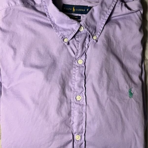 Ljuslila skjorta från Ralph Lauren - Snygg ljuslila skjorta från Ralph Lauren med klassisk krage och knappar framtill. Skjortan har en broderad logga i grönt på bröstet. Perfekt för en stilren look!