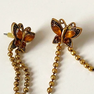 Guldiga fjärilsörhängen med kedjor - Snygga guldiga örhängen formade som fjärilar med långa kedjor. Fjärilarna har detaljer i orange och små pärlor som ger en elegant touch. Perfekta för att ge en unik stil till din outfit.