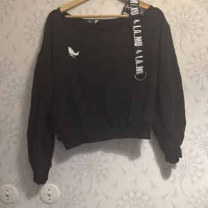 Svart offshoulder tröja med banddetalj - Snygg svart offshoulder tröja från Lamu med en cool banddetalj på axeln. Tröjan har långa ärmar och en avslappnad passform, perfekt för en trendig look. Passar bra till både jeans och kjol!