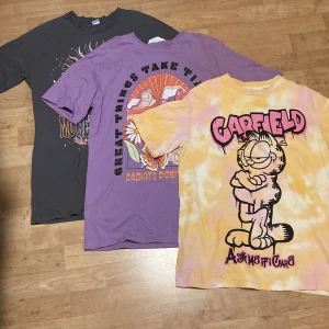 Tre färgglada t-shirts med tryck - Säljer tre coola oversized t-shirts med olika tryck. En gul och rosa tie-dye med Garfield-motiv, en lila med blommigt tryck och texten 'Great Things Take Time to Grow', och en mörkgrå med sol- och månmotiv och texten 'Mother Universe'. Perfekta för en avslappnad stil!