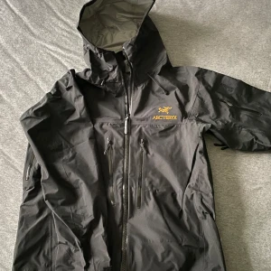 Svart skaljacka från Arc'teryx - Säljer en svart skaljacka från Arc'teryx i GORE-TEX Pro-material. Jackan har en justerbar huva och flera praktiska fickor med dragkedjor. Perfekt för utomhusaktiviteter och skyddar mot väder och vind.