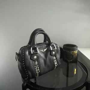 Svart handväska från Zadig & Voltaire - Snygg svart handväska i skinn från Zadig & Voltaire i den minsta storleken ”Sunny Nano”⚡️⚡️Den är i använt skick inga defekter endast av färgningar på silvret som man ser på bilderna🥰 Väskan medföljer ett avtagbart axelband där det står ”Voltaire” samt får du med dustbag som tillhör väskan💕🌸 