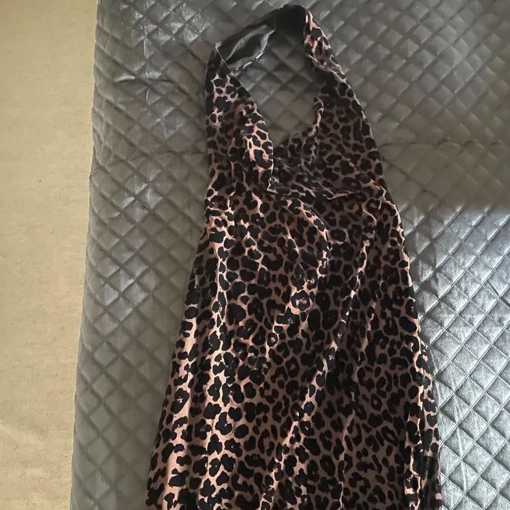 Snygg leopardmönstrad långklänning med djup v-ringning och halterneck-design. Perfekt för att sticka ut med stil. Klänningen har en figurnära passform som framhäver silhuetten.. Mekot.