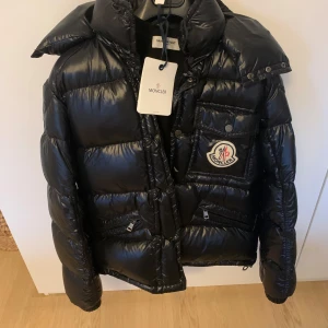 Svart dunjacka från Moncler - Säljer en snygg svart dunjacka från Moncler med glansig finish. Jackan har en stor logotyp på bröstfickan och flera praktiska fickor med dragkedja. Perfekt för kalla dagar med sin varma dunstoppning. Jackan har långa ärmar och en hög krage för extra skydd mot kyla.storlek 2