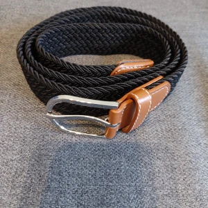 Svart flätat bälte med bruna detaljer - Snyggt svart flätat bälte med bruna läderdetaljer och en stilren silverfärgad spänne. Perfekt för att ge en extra touch till din outfit. Bältet har en klassisk design med kontrasterande sömmar på läderdetaljerna.