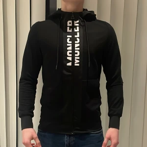 Svart hoodie från Moncler - Snygg svart hoodie från Moncler med dragkedja och justerbar huva. Moncler-logga på ärmen och stort tryck på framsidan Ny pris 2200kr, använd endast 2 gånger! Riktigt stilren och fräsch hoodie. Mega otroligt bra pris!!✅