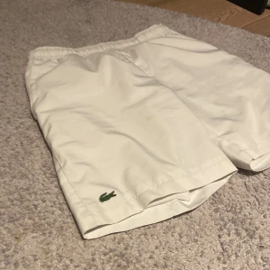 Vita sportshorts från Lacoste - Säljer ett par vita sportshorts från Lacoste med elastisk midja och den klassiska krokodilloggan. Perfekta för en aktiv dag eller bara för att chilla. De är gjorda i ett lätt och bekvämt material.