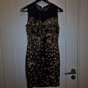 Svart Cocktailklänning med Guldbroderi - Säljer denna eleganta klänning i storlek 38-40. Klänningen är i svart med vackra guldbroderade detaljer, perfekt för festliga tillfällen eller en utekväll. Den sitter bekvämt och har en dragkedja baktill för enkel på- och avklädning. Säljer för 499:-