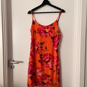 Orange blommig klänning - Säljer en superfin orange klänning med stora blommor i rött och rosa. Den är perfekt för sommaren med sina tunna axelband och lätta material. Klänningen är i storlek L och har en härlig, färgglad design som verkligen sticker ut. 
