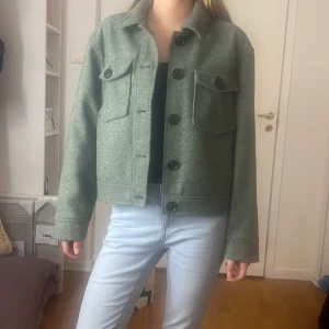 Grön jacka med stora knappar - Säljer en snygg grön jacka med stora knappar framtill. Den har två stora fickor på bröstet och en rak passform. Perfekt för höst och vår, och den är superbekväm med långa ärmar. Jackan är i bra skick och passar till både jeans och kjol.