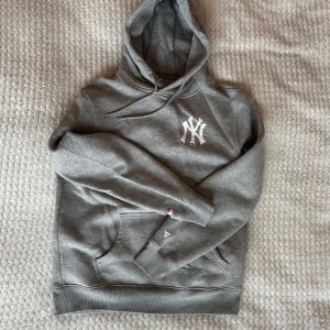 Grå hoodie från New York Yankees - Säljer en grå hoodie från New York Yankees i storlek XL. Den har en stor logga på framsidan och flera tryck på baksidan med Yankees-tema. Perfekt för höst och vinter, och den är superbekväm med en känguruficka och justerbar huva. Nytt skick, aldrig använd. Går liten i storlek, passar mer som M/L.