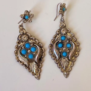 Vintageinspirerade örhängen med turkosa stenar - Säljer ett par unika vintageinspirerade örhängen i silverfärg med vackra turkosa stenar. De har en elegant, droppformad design med blommönster och små detaljer som ger en bohemisk känsla. Perfekta för att ge en färgklick till din outfit!