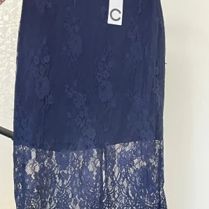 Mörkblå spetskjol med dragkedja - Säljer en superfin mörkblå spetskjol som är perfekt för både fest och sommar. Kjolen har en elegant dragkedja baktill och ett vackert blommigt mönster. Den är i nyskick och har en stilren design med en längre spetsdetalj längst ner. Passar perfekt till en somrig topp eller en festlig blus!