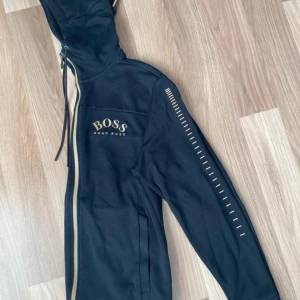 Svart jacka från Hugo Boss - Säljer en snygg svart jacka från Hugo Boss. Den har en dragkedja framtill och en huva med snören. Ärmarna är långärmade med coola detaljer längs sidorna. Perfekt för höst och vår!