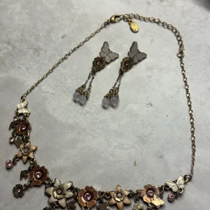 Blommigt halsband och örhängen med fjärilar - Säljer ett superfint set med halsband och matchande örhängen. Halsbandet har en kedja i antik guldton och är dekorerat med blommor i olika nyanser av brunt och beige, samt små rosa stenar. Örhängena har en söt fjäril högst upp och hängande blommor med liknande rosa stenar. Perfekt för att ge en bohemisk touch till din outfit!