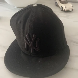 Svart keps från New Era - Säljer en svart keps från New Era med New York Yankees-loggan framtill. Kepsen är i bra skick och har en klassisk design med böjd skärm. Perfekt för vardagsbruk eller sportevenemang.